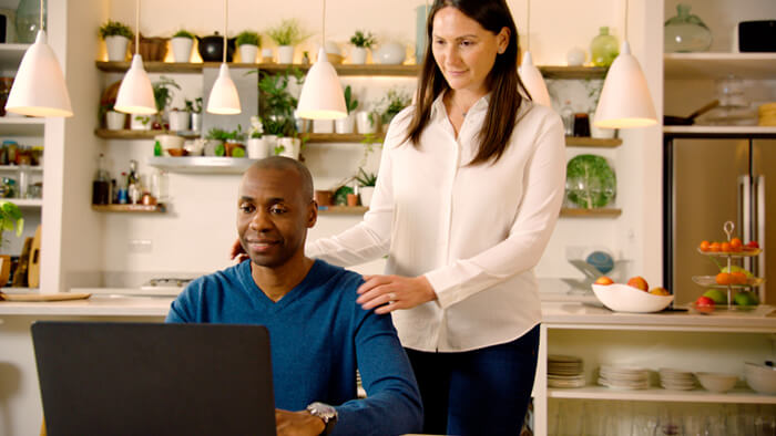 Mies istuu keitti&ouml;ss&auml;, ja vaimo seisoo h&auml;nen vieress&auml;&auml;n. Molemmat katsovat kannettavaa tietokonetta.