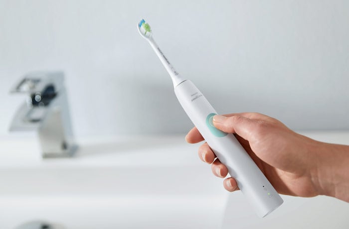  Joku pitää valkoista Philips Sonicare -sähköhammasharjaa käsissään lavuaarin ääressä sormi painikkeella.