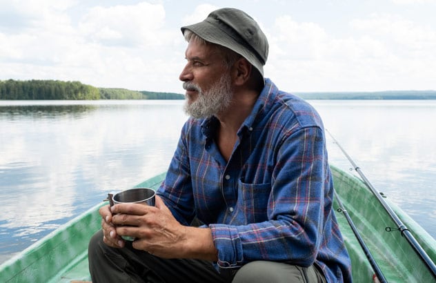 Vanhempi mies, jolla on harmaa parta, kalastajanhattu ja siniruutuinen paita, istuu veneessä ja katsoo kaukaisuuteen.