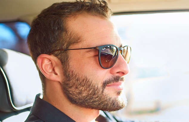 Mies ajaa autoa mustat aurinkolasit päässä. Hänellä on täyspitkä ruskea parta, viikset sekä lyhyet hiukset.