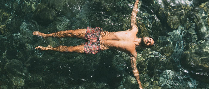 Laajakuva parrakkaasta miehestä uimashortseissa kellumassa kasvot ylöspäin vedessä käsivarret sivuille levitettynä.