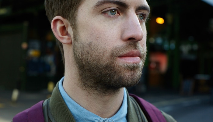 Ruskeahiuksinen mies, jonka kasvoja ja kaulaa peittää 1 cm:n pituinen parta, seisoo kadulla violetti selkäreppu selässä.